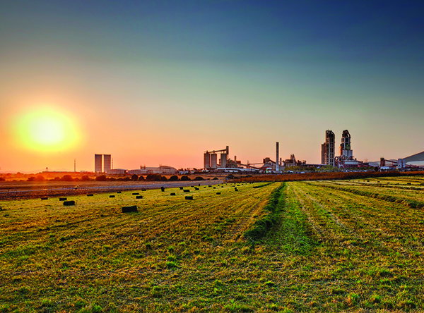 A cement plant on the horizon beside a grassy field at sunset, with smokestacks and industrial buildings silhouetted against the sky.