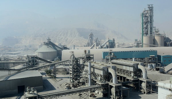Industrial cement plant with multiple conveyors, silos and processing buildings, seen against a hazy mountainous background.