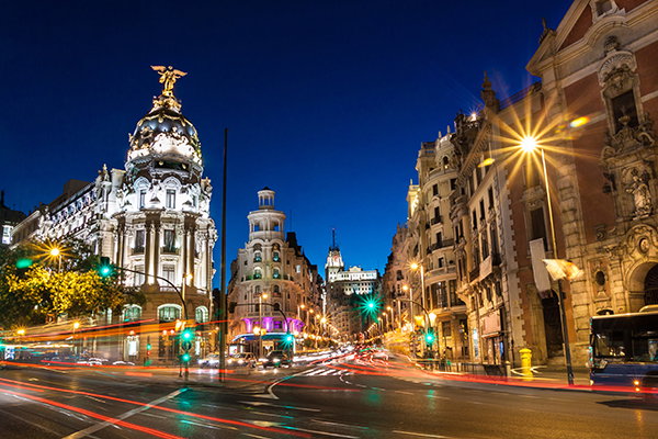 Gran Via, Madrid, Spain - nighttime city street with illuminated historic buildings, a domed landmark, and long-exposure light trails from passing traffic.
