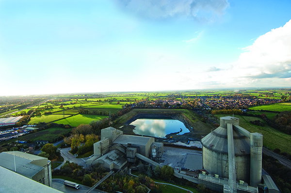 Aerial view of a cement plant with large silos and industrial buildings beside a pond, surrounded by green fields and countryside under a cloudy sky.