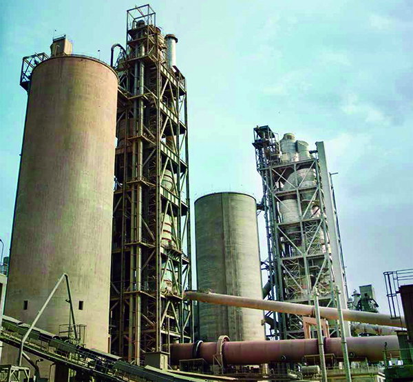 Cement plant in Pakistan with large storage silos, metal towers and pipes against a pale sky.