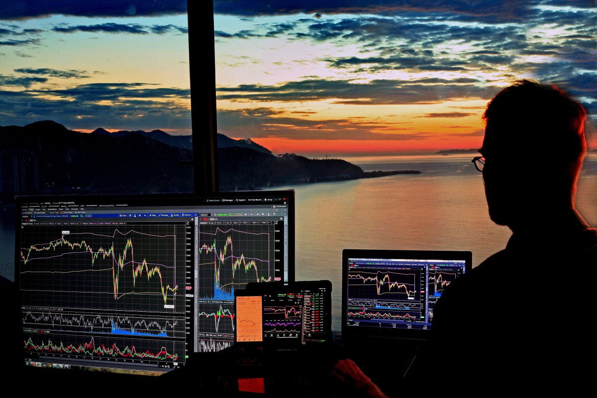 Silhouette of a person at a desk looking at multiple monitors filled with market charts, with a sunset over water and mountains outside the window.