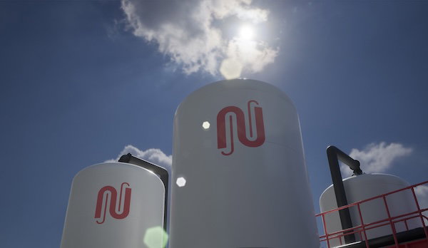 Large white industrial tanks with a red logo, seen against a blue sky with bright clouds and sun glare.