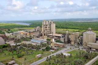 Wide view of a large cement plant with silos, industrial buildings and surrounding green landscape under a cloudy sky.