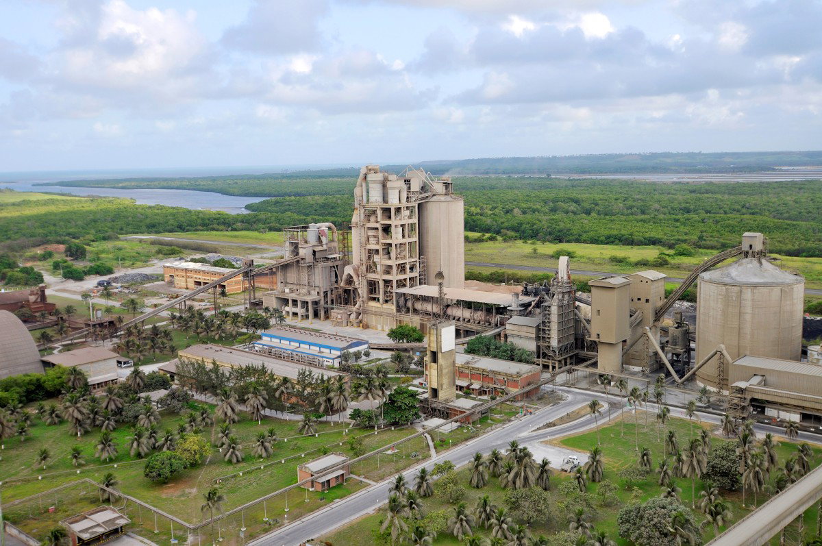Wide view of a large cement plant with silos, industrial buildings and surrounding green landscape under a cloudy sky.