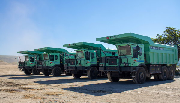 Three large green haul trucks are parked side by side on a dusty quarry road under a clear sky.