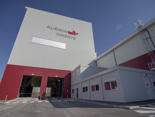 Exterior of a large industrial cement plant building with white and red cladding, loading bays and small windows, seen under a clear blue sky.