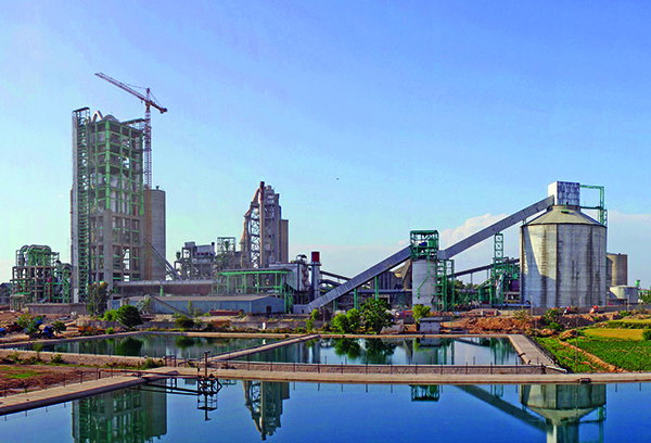 Cement plant with tall silos, a conveyor bridge and a crane beside a water channel, with landscaped grounds in the foreground