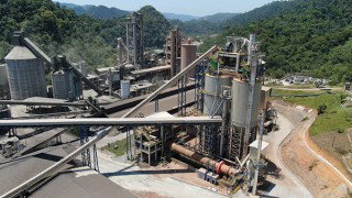 A cement plant with silos, conveyors and industrial structures set among green hills and forested mountains.