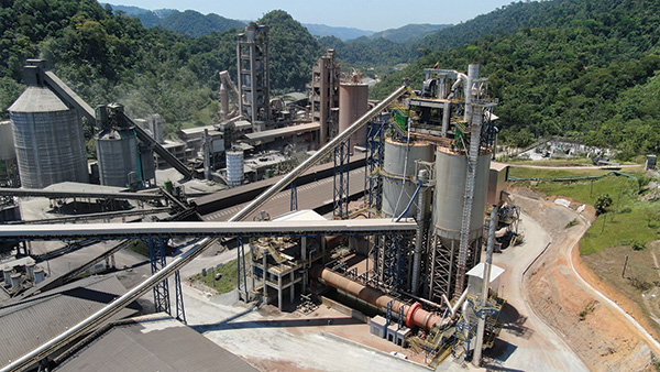 A cement plant with silos, conveyors and industrial structures set among green hills and forested mountains.
