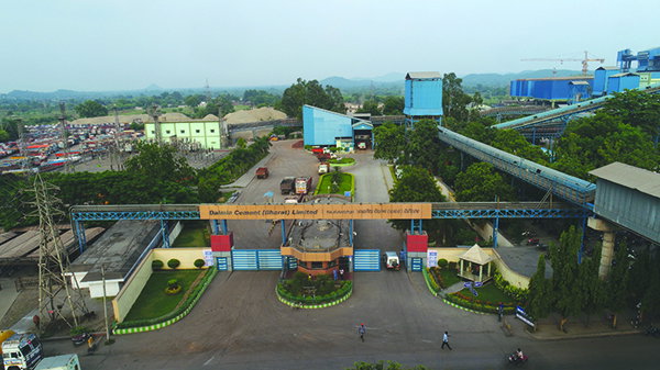 Wide view of a cement plant entrance with an overhead conveyor, factory buildings and storage areas in the background, seen from above.