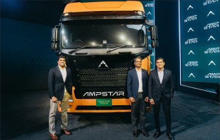 Three men stand in front of a large orange-and-black electric truck displayed at an event, with branding visible on the vehicle and backdrop.