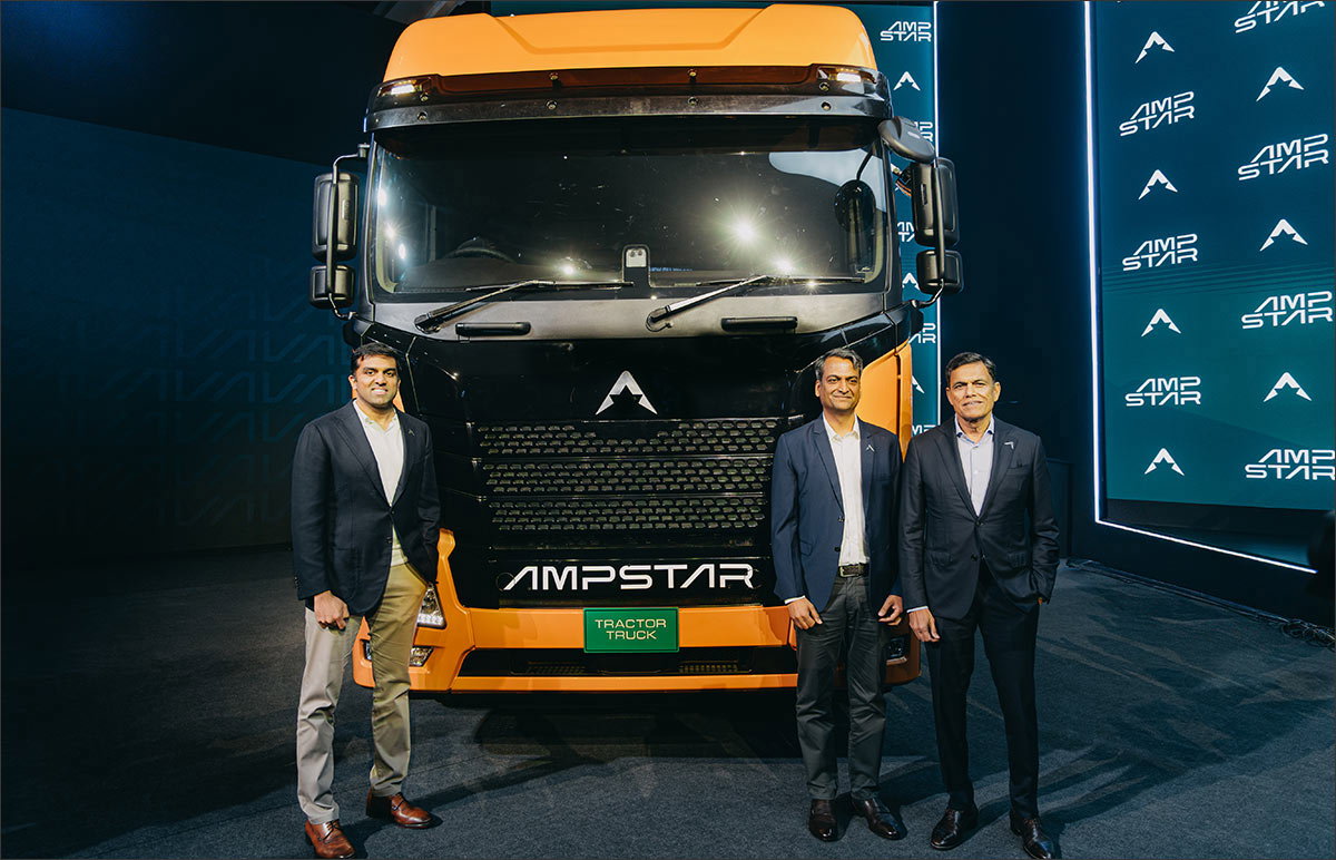 Three men stand in front of a large orange-and-black electric truck displayed at an event, with branding visible on the vehicle and backdrop.