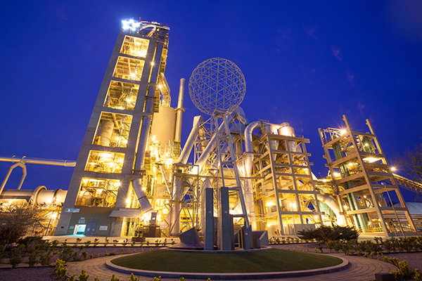 Night view of a brightly lit industrial plant with tall towers, pipes and illuminated structures against a dark sky.