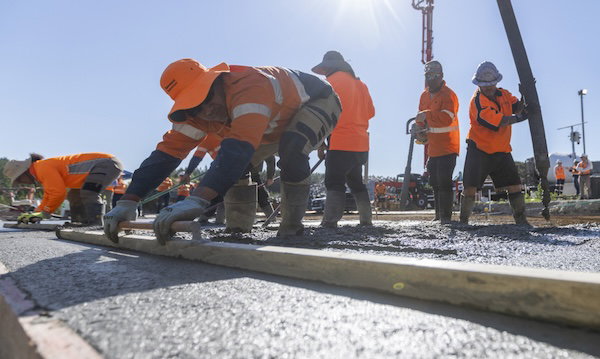 Workers in orange safety gear lean over fresh concrete along a road edge at a construction site in bright sunlight.