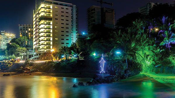 Nighttime cityscape of Dakar, Senegal, with a lit high-rise building, illuminated trees, and colorful reflections on the water in the foreground.