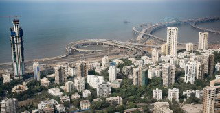 A coastal city skyline with high-rise buildings beside sweeping highway interchanges and bridges over the water.