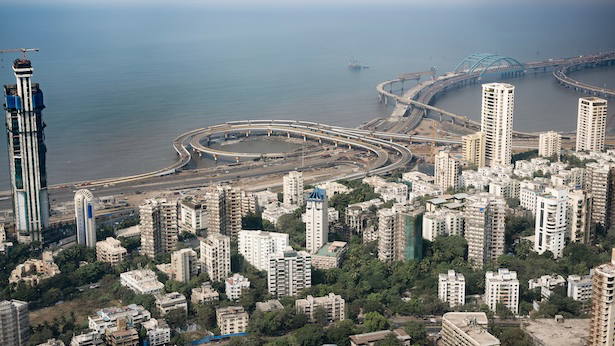 A coastal city skyline with high-rise buildings beside sweeping highway interchanges and bridges over the water.