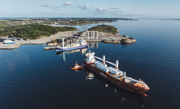 Aerial view of two cargo ships moored beside a coastal port, with tugboats nearby and open sea stretching to the horizon.
