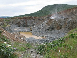 A rocky open pit quarry with muddy water pooled at the bottom and green vegetation around the edges.