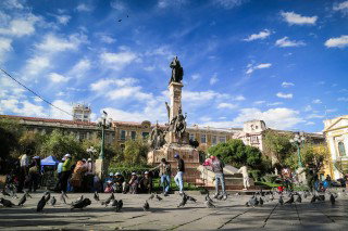 A busy city square in La Paz, Bolivia, with a tall monument in the centre, people walking and gathering around it, and buildings under a blue sky with scattered clouds.