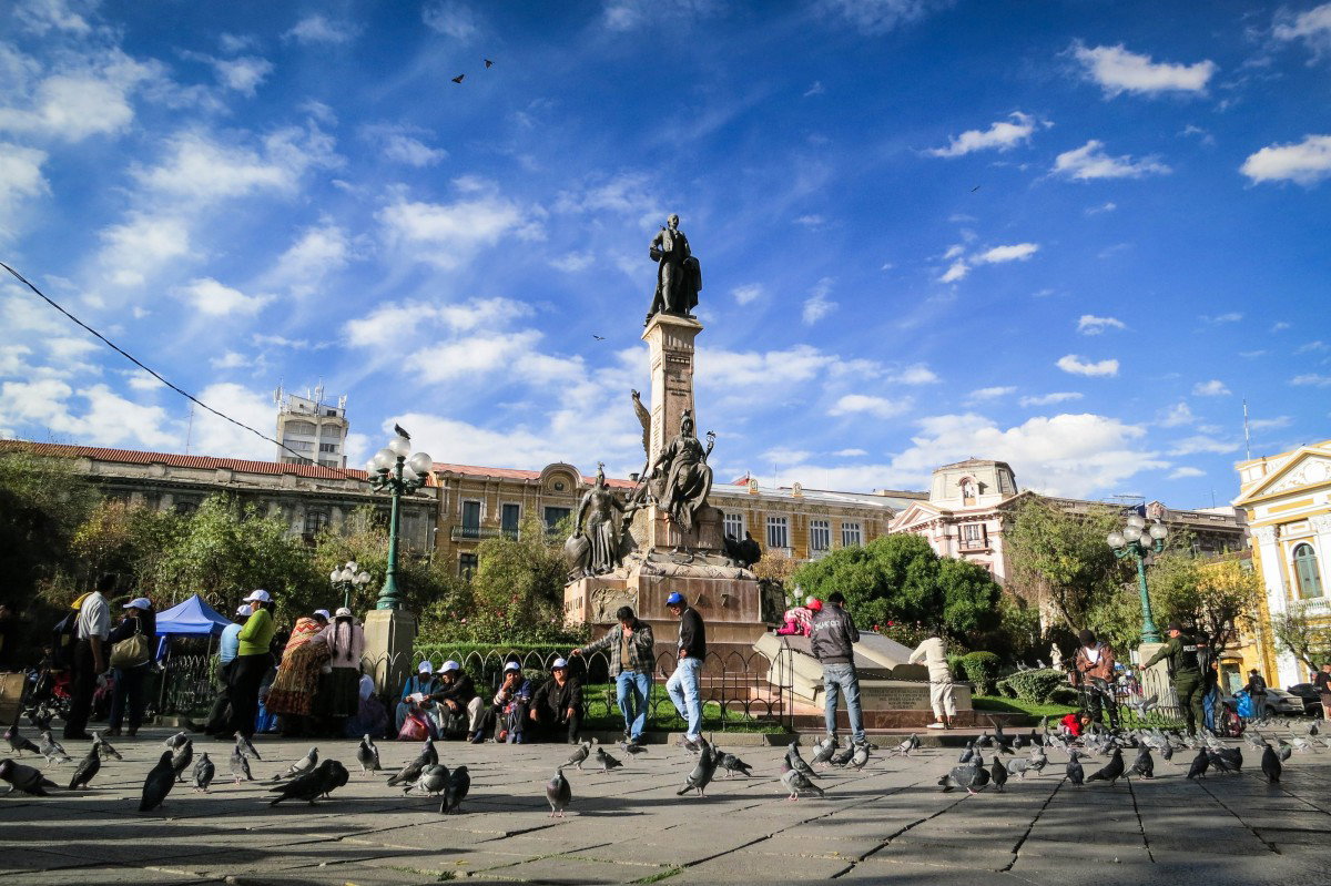 A busy city square in La Paz, Bolivia, with a tall monument in the centre, people walking and gathering around it, and buildings under a blue sky with scattered clouds.