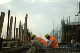 Two construction workers in hard hats and high-visibility vests crouch over steel rebar on a building site, with formwork and scaffolding in the background.