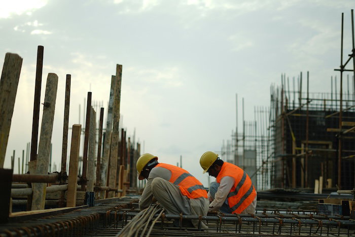 Two construction workers in hard hats and high-visibility vests crouch over steel rebar on a building site, with formwork and scaffolding in the background.