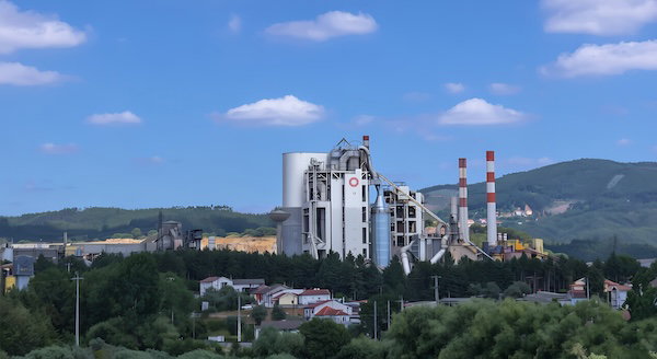 A large cement plant with tall silos and smokestacks stands beside green hills under a blue sky.